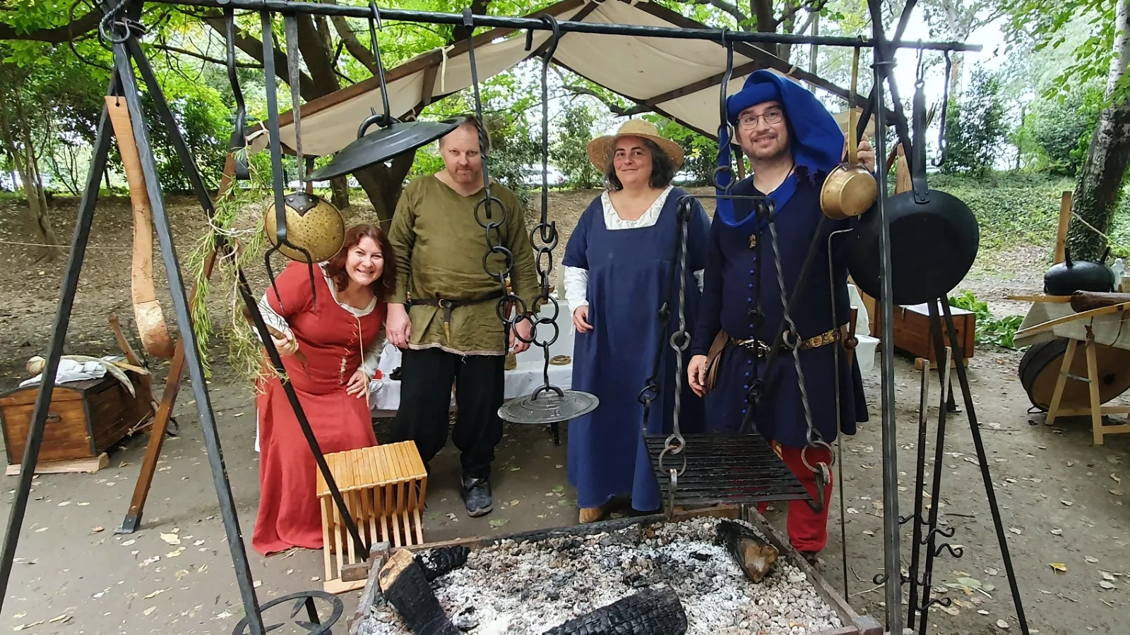 Quatre membres de la troupe en costume, souriants, derrière la table à feu et ses ustensiles suspendus, sous l’auvent du camp